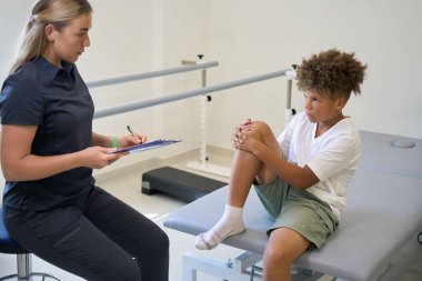 Terapist not alırken hasta terapi masasında oturuyor. Muhtemelen son yapılan bir değerlendirmenin sonuçlarını ya da gözlemlerini kaydediyor.