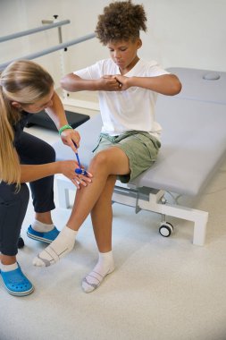 Female physical therapist tests the muscle reflexes of a teenage boy using a special instrument clipart