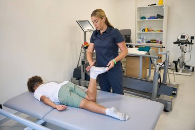 Terapist bacak hareketliliğini ve kas esnekliğini değerlendirirken hasta terapi masasında yatıyor.