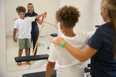 Uzman, genç bir hastaya omuz ve kol kaslarını güçlendirmek amacıyla direnç bandı egzersizi yapmasına yardım ediyor.