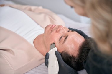 Rahatlamış bir adam, ultrasonik temizleme işlemi sırasında tedavi yatağında yatar. Yüzü daha taze ve yumuşaktır. Gözeneklerini temizler ve cildinin berraklığını arttırır.