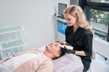 Gülümseyen kadın kozmetik uzmanı, erkek hasta tenindeki kirliliği ve fazlalığı gidermek için ultrasonik fırça kullanıyor.