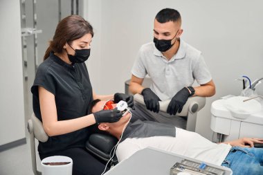 Profesyonel dişçi ekibi diş beyazlatma prosedürü uyguluyor, erkek hasta dişlerine mor ötesi ışıkla özel bir cihaz uyguluyor, müşteri UV koruyucu gözlüklerle diş sandalyesinde yatıyor.