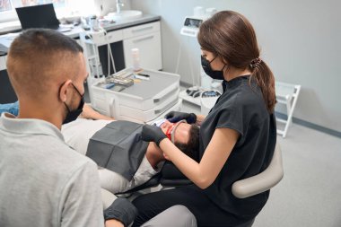 Erkek dişçi asistanı, dişlerini beyazlatma prosedürünü koruyan yüksek nitelikli dişçiyi izliyor. Ultraviyole ışıklı, estetik dişli özel bir cihaz kullanıyor.