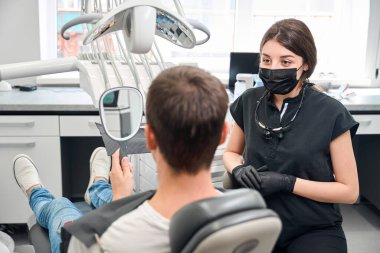 Back view male client of aesthetic dentistry sitting on dental chair and looking at the mirror at his healthy white teeth, qualified woman dentist sitting nearby clipart