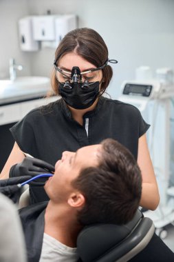 Üniformalı nitelikli kadın dişçi ve özel gözlükler erkek müşteriye yüksek nitelikli diş tedavisi sağlıyor, diş kökleriyle ve kanallarla çalışıyor, özel pulluk kullanarak ceplerini temizliyor.