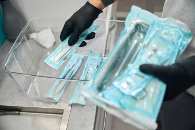 Lastik eldivenli diş teknisyeni paketleri depodan steril aletlerle alıyor, ofisi işe hazırlıyor, hastaları bekliyor, sağlık hizmetlerine karşı sorumlu tutum sergiliyor.