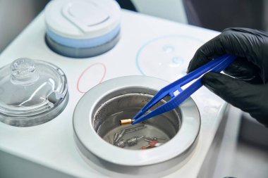 Lastik eldivenli diş teknisyeni cımbızla diş hortumunu tutuyor ve ultrasonik banyoya sokuyor, yüksek bir tıbbi temizlik ve dezenfeksiyon standardı elde ediyor.