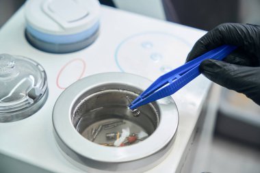 Diş hekimi diş hortumlarını ultrasonik otomatik temizleyiciye koyar. Cımbız kullanır, diş aletlerinin etkili temizliği, güvenlik ve hijyen sağlar.