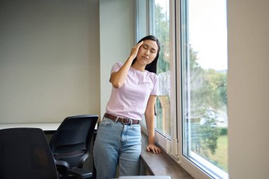 Genç bir kadın iş yerinde baş ağrısı çekiyor, büyük bir pencerenin yanında duruyor.