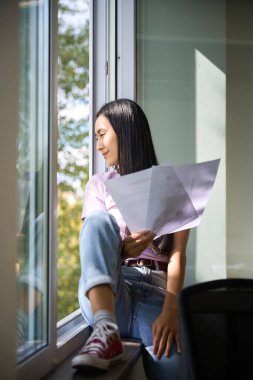 Kadın serbest çalışan geniş bir pencerenin pencere eşiğinde proje belgeleriyle oturuyor, rahat kıyafetler giyiyor.