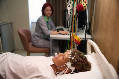 Sıhhiyeci, hasta yatakta yatarken monitörde EEG okumalarını gözlemler ve nörolojik test sırasında doğru veri toplamasını sağlar.