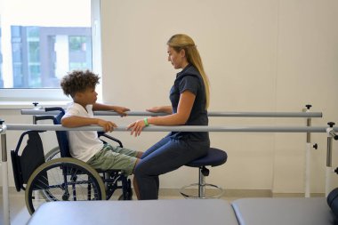 Terapist, ayakta durma ve dengeleme egzersizlerini paralel çubuklarla yaparken hastayı teşvik eder ve motive eder.