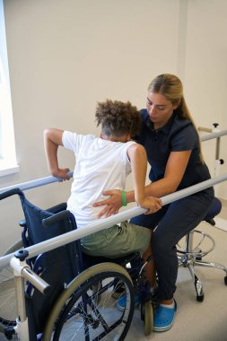 Fizyoterapist, tekerlekli sandalyeden ayakta durmaya geçerken hastaya denge kurma pratiği yaparken destek sağlar.