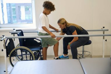 Terapist, genç bir hastaya tekerlekli sandalyedeki pozisyonundan paralel parmaklıkların desteğiyle ayakta durabilmesi için yardımcı oluyor.