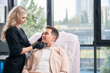 Siyah üniformalı ve siyah lastik eldivenli çekici kadın bej takım elbiseli yetişkin erkek müşterinin derisini dikkatlice inceliyor, estetik erkek yüzü temizliyor.