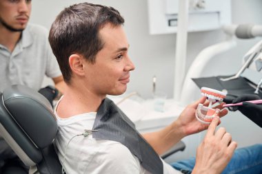 Dişçilik bürosunun erkek müşterisi sandalyeye oturmuş elinde çenesinin 3D modelini tutuyor ve doktora dişlerinin sorunlarını soruyor.