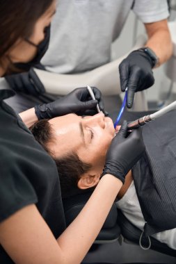 Yandan bakan profesyonel diş hekimi ekibi erkek dişlerini tedavi ediyor, diş çürümesini ve diş minesi tahribatını önlemek için dolgu yapıyor ve yerleştiriyor, estetik dişçilik.