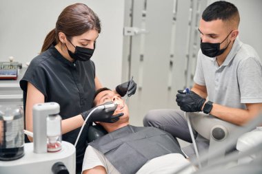 Farklı aletlerle evrensel ölçeklendirici kullanan deneyimli bir dişçi ve hasta erkek dişlerini tedavi eden hortumlar, ona yardım eden erkek asistan, estetik dişçilik.