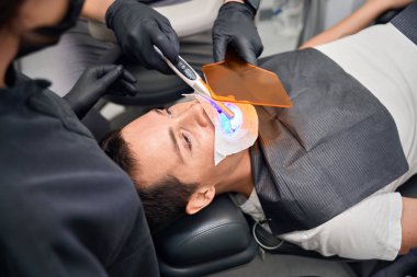 Diş beyazlatma prosedürüne katılan genç bir adam, dişçi koltuğunda yatarken yetenekli bir doktor UV lambası ve özel beyazlatma maddesi, estetik dişçilik uygulaması yapıyor.