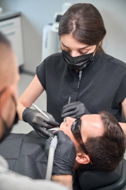 Arkadan bakan dişçi asistanı elinde salya püskürtücü tutarken, kadın doktor da erkek hasta ağzında sorunlu dişlerle, profesyonel bakım ve bakım yaklaşımıyla çalışıyor.