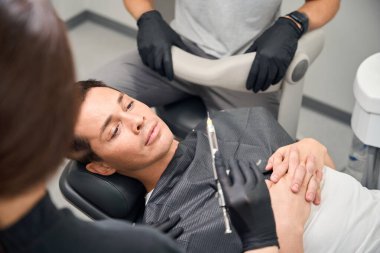 Estetik dişçilik kliniğinin korkmuş erkek müşterisi büyük anestezik diş şırıngasına bakıyor, enjeksiyondan önce korku hissediyor.