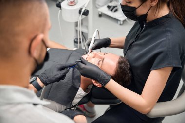 Arkaya bakan erkek dişçi, ağız boşluğu sağlığını kontrol etmek ve hastanın dişlerini temizlemek için ultrasonik pulu kullanan deneyimli kadın doktora yardım ediyor.