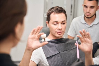 Kadın ve erkek dişçiler diş implantı yerleştirme sürecini müşterilerine açıklıyorlar. Ellerinde büyümüş 3 boyutlu implant ve diş protezi tutan adam doktorları dinliyor.