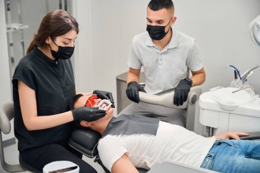 Deneyimli stomatolog, diş sandalyesinde koruyucu gözlükleriyle yatan, diş beyazlatma prosedürü için stomatolojiste giden erkek müşteriye ağız açıcı ayarlıyor.