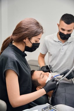 Diş sandalyesinde yatan erkek hastalara yüksek kaliteli diş bakımı sağlayan koruyucu yüz maskesi uzmanlarına bakın.