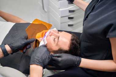 Diş asistanının elinde özel UV lambası ve koruyucu cam var. Sandalyede yatan erkek müşteriye diş beyazlatma prosedürü uygulanıyor.