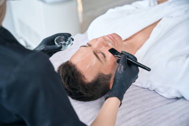 Bir kozmetolojistin kaplıca merkezinde genç bir adamın yüzüne maske takmasının üst görüntüsü.
