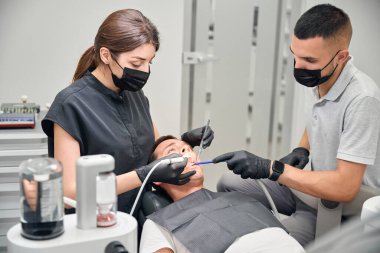 Diş hekimi asistanı elinde tükürük çıkarıcı tutarken kadın doktor da erkek dişlerini ultrasonik pullukla tedavi ediyor.