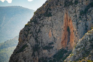 Parlak gün ışığı altında sakin dağlık bölgede görkemli kaya oluşumu yükseliyor. Sakin atmosfer doğanın keşfine davet ediyor güzellik ve ihtişam.
