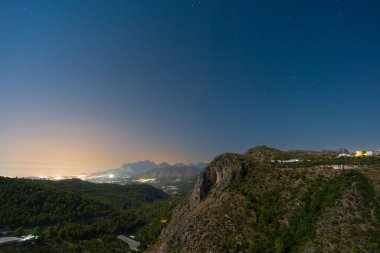 Huzurlu gece manzarası görkemli dağlar ve uzak şehir ışıklarıyla aydınlatılmış bir vadi. Yıldızlı gökyüzü manzaranın huzurlu atmosferine katkı yapıyor..