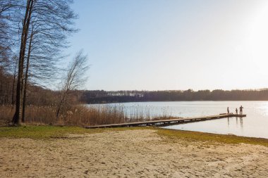 Güneşli bir günde ağaçlarla, rıhtımlarla ve balıkçıların siluetleriyle dolu bir göl sahili.