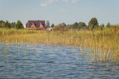 Lake on a sunny day with ripples on the surface of the water, blue sky and white clouds. View from the water, to the shore cottages. clipart