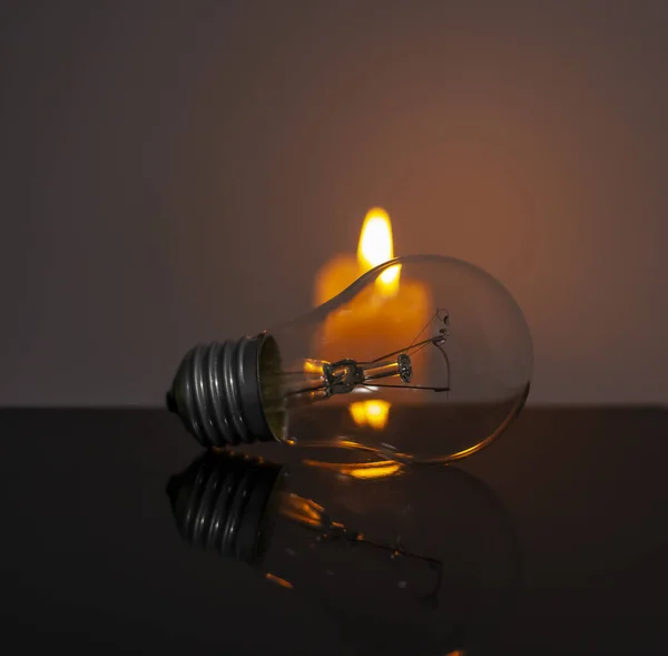 Burning candle near a switched off light bulb in complete darkness. Blackout, electricity off, energy crisis or power outage, concept image.