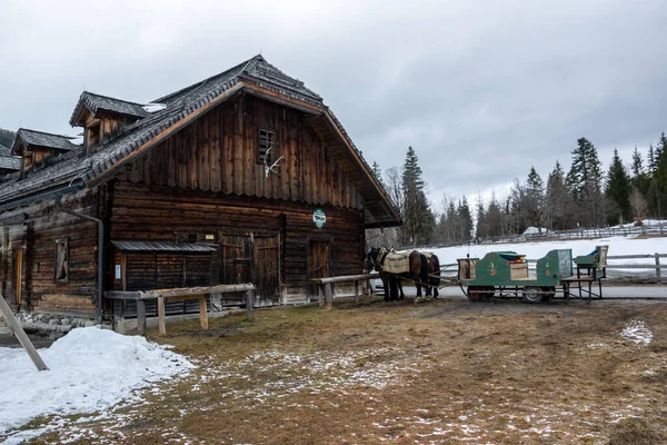 A farm with a wooden barn in winter in Austria. Horses with a carriage in front of the stable.