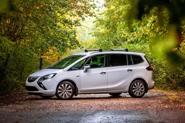 Sonbaharda ormanda ağaçların arasında beyaz aile arabası MPV Opel Zafira. Sonbahar yaprakları yere düşüyor, ağaçlarda yeşil ve renkli yapraklar..