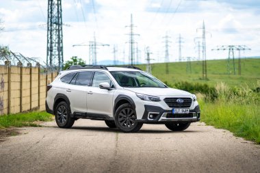 Brno, Çek Cumhuriyeti - 2 Haziran 2024: Beyaz, modern bir Subaru Outback SUV arka planda elektrik direkleri olan eski bir elektrik santrali duvarının önünde park halinde..
