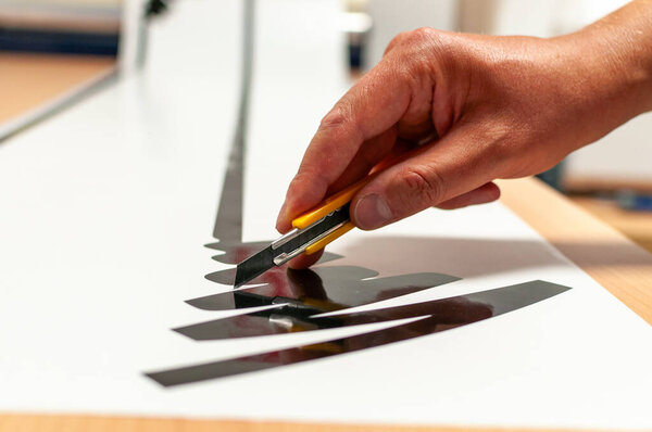 The hand of a man at work holding a knife and making advertising stickers in cut foil.