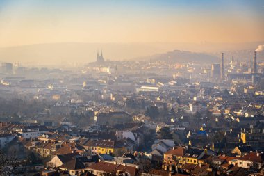 Brno, Çek Cumhuriyeti - 10 Ocak 2025: Gün batımında şehrin bulutlu ve sisli manzarası. Şehrin üzerindeki gökyüzü güneş ışığıyla yıkanır ve evler kış havasının tersine döndüğünde puslu olur..