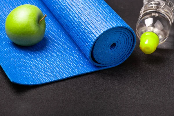 Blue yoga mat, water bottle, apple on dark background. Equipment for yoga. Healthy lifestyle, sport, diet, meditation, relaxation. Workout at home or gym. Fitness yoga and exercise concept with copy spac