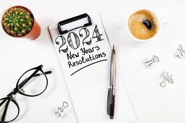 New year resolutions 2024 on desk. 2024 goals list with notebook, coffee cup, plant on white table. Resolutions, plan, goals, action, checklist, idea concept. New Year 2024 resolutions, copy space