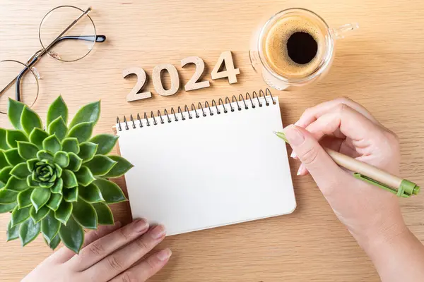 New year resolutions 2024 on desk. 2024 goals - hands and notebook, coffee cup, plant on wooden table. Resolutions, plan, goals, action,  idea concept. New Year 2024 resolutions, copy space