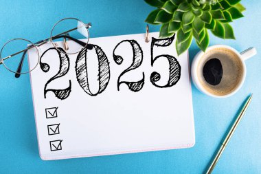 Yeni yıl kararları 2025 masada. 2025 gol listesi, defter, kahve fincanı, mavi masaya bitki. Kararlar, plan, hedefler, eylem, kontrol listesi, fikir konsepti. Yeni yıl 2025 kararları. Boşluğu kopyala