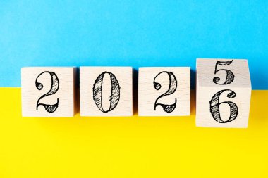 2026 Mutlu yıllar konsepti. Sarı ve mavi arkaplan Ukrayna bayrağında 2025 'ten 2026' ya tahta blok değişimi. Siyaset, Ukrayna 'ya destek, çözümler, hedefler, iş dünyası, başarı fikirlerine ilham