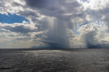 Rainfall over the sea with dramatic clouds, sunlight reflections, and distant mountains on the horizon in summer clipart