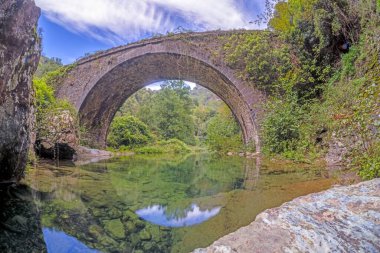 Korsika 'daki tarihi taş köprü Ponte di Infernu, gündüz vakti bereketli doğayla çevrili berrak sulara yansıyor.